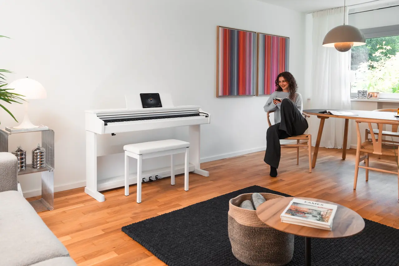 Piano numérique Kawai blanc dans un salon lumineux ; au fond, une femme est assise à table.